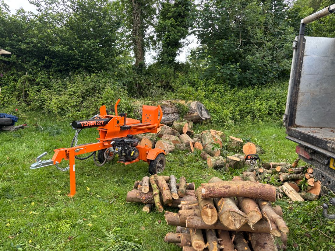 A towable hydraulic log splitter set up on grass beside cut rounds, split logs and a stack of cordwood, with the tipper truck alongside